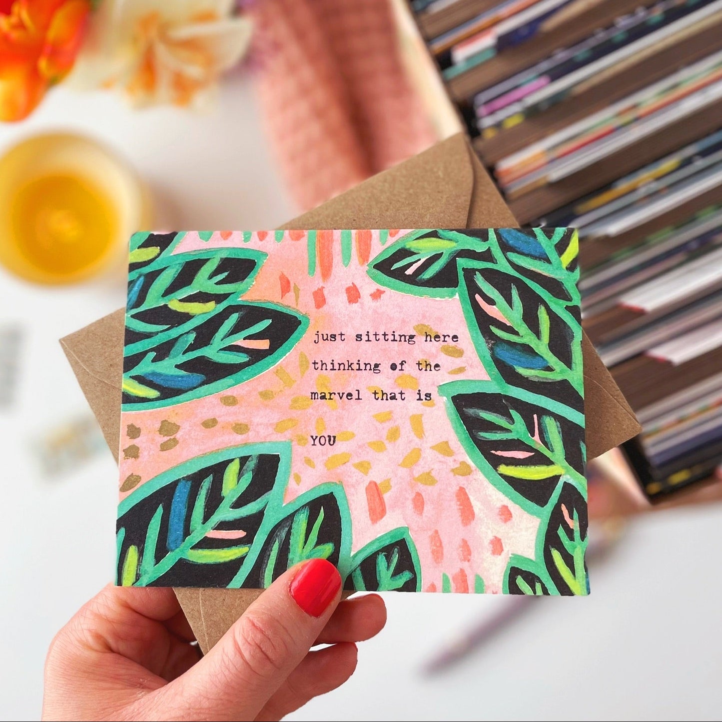 Hand holding a colorful card with leaf design and text, surrounded by flowers and books.