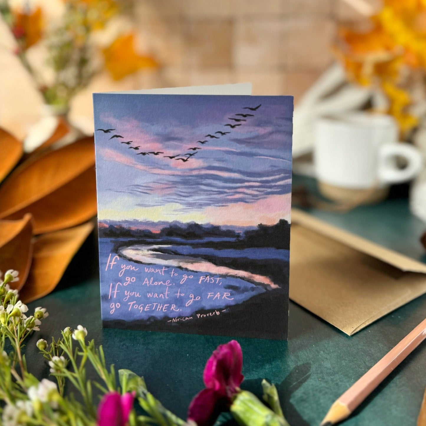 Greeting card with a scenic design of birds flying over water, placed on a table with flowers and a pencil.