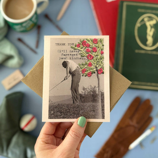 Hand holding a vintage-style card with a golfing scene and tree, surrounded by books and stationery on a blue surface.