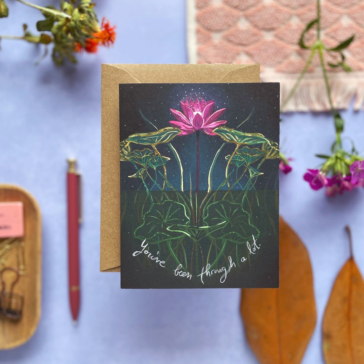 A greeting card with a kraft envelope, featuring an illustration of a pink lotus flower on dark green leaves against a black background, with the handwritten text 'You've been through a lot' at the bottom. The card is styled on a light blue surface with dried orange flowers, a red pen, a wooden tray with paperclips, and a pink patterned fabric swatch.