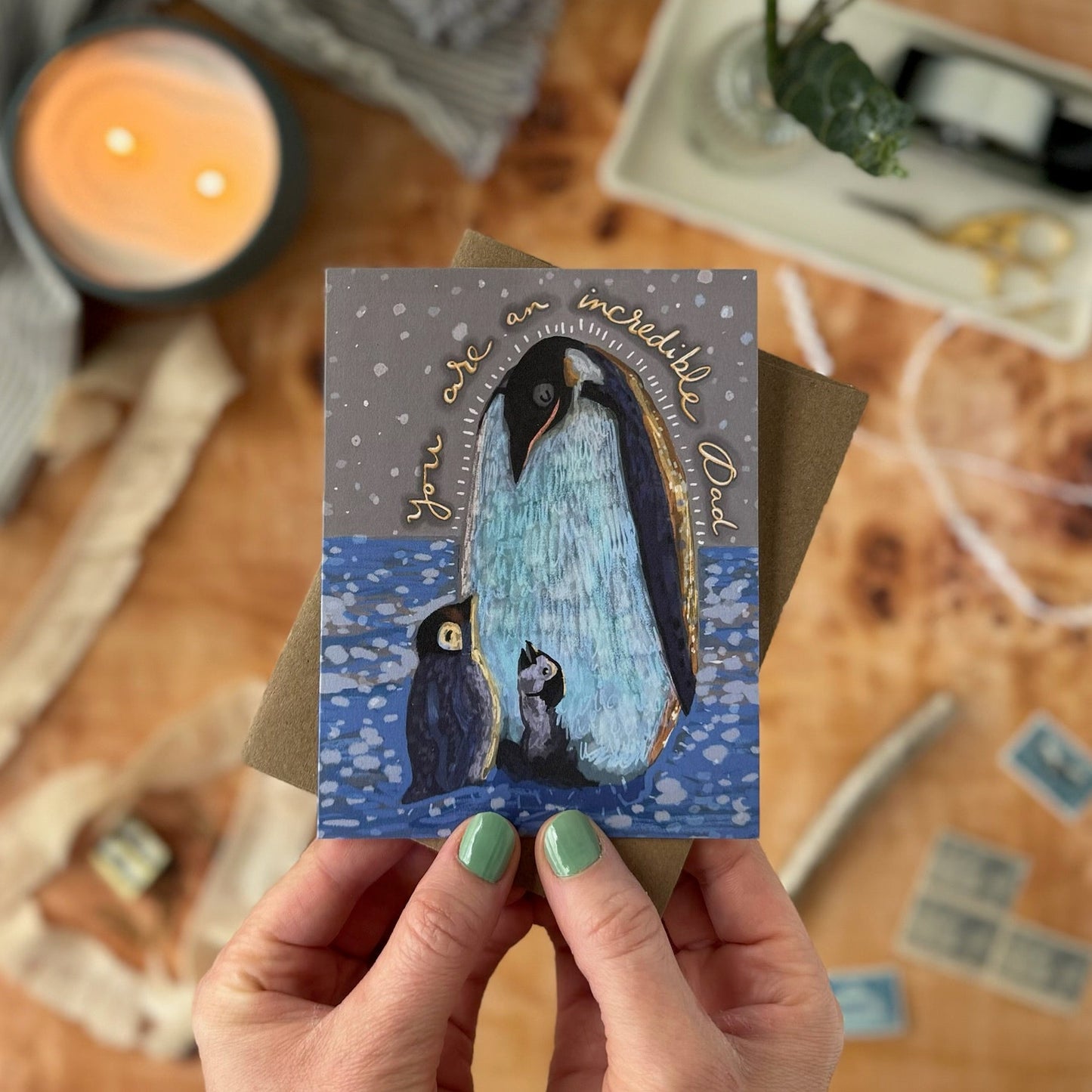 Hand holding a small artwork of a daddy penguin and two chicks, with a snowy landscape and text, on a wooden surface with a candle and flowers.