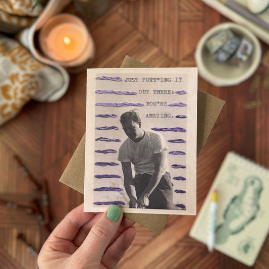 Hand holding a card with text and a person's silhouette on a wooden surface with candles and plants.