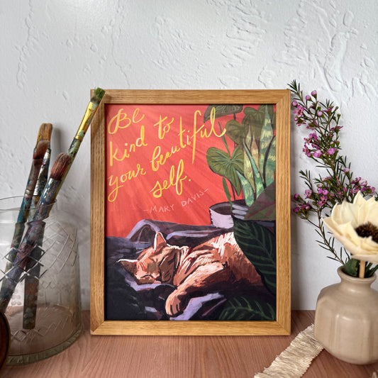 A framed art print featuring a sleeping orange cat on a red background with the quote 'Be kind to your beautiful self. - Mary Davis' in gold lettering.