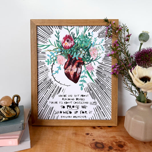 A framed art print featuring a watercolor anatomical heart surrounded by pink and red flowers and green leaves, with black hand-drawn rays and text, displayed on a wooden table with flowers and books.