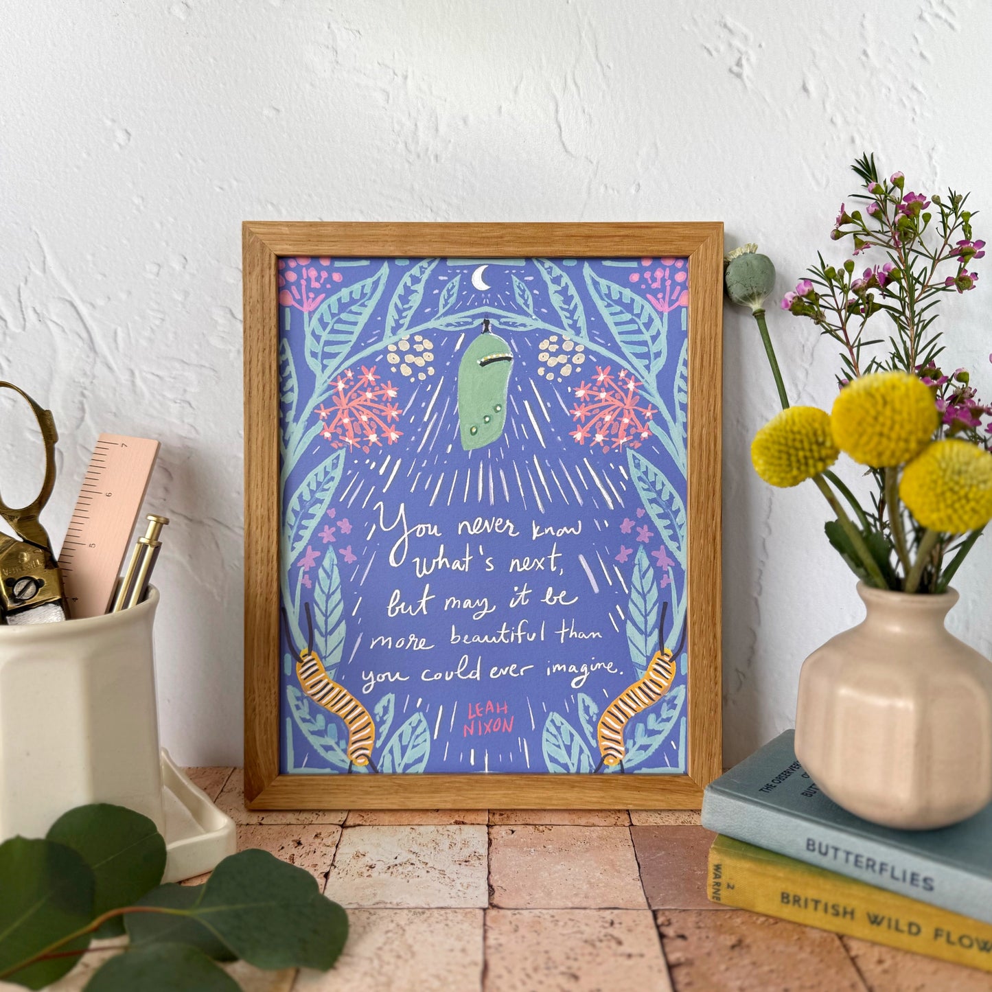 A framed art print featuring a whimsical purple and blue illustration with a green chrysalis, caterpillars, flowers, and handwritten text, styled on a terracotta tile surface next to a vase of yellow flowers and desk accessories.