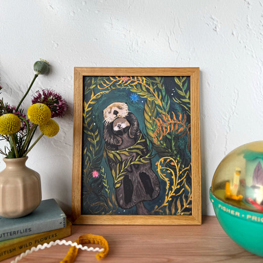 A framed art print featuring an illustration of a sea otter mother holding her baby, nestled among lush green and yellow foliage, styled on a wooden tabletop next to a vase of flowers and a green children's toy ball against a white textured wall.