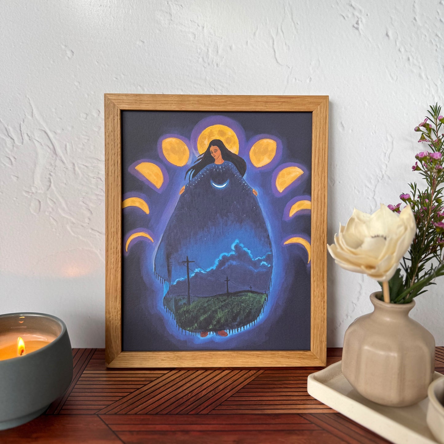 A framed art print featuring a surreal night scene with a glowing form of a woman surrounded by moon phases, displayed on a wooden table against a textured white wall with a candle and flowers nearby.