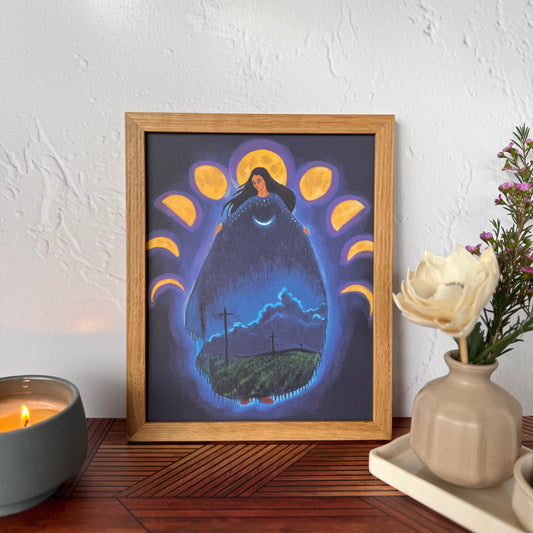 A framed art print featuring a surreal night scene with a glowing form of a woman surrounded by moon phases, displayed on a wooden table against a textured white wall with a candle and flowers nearby.