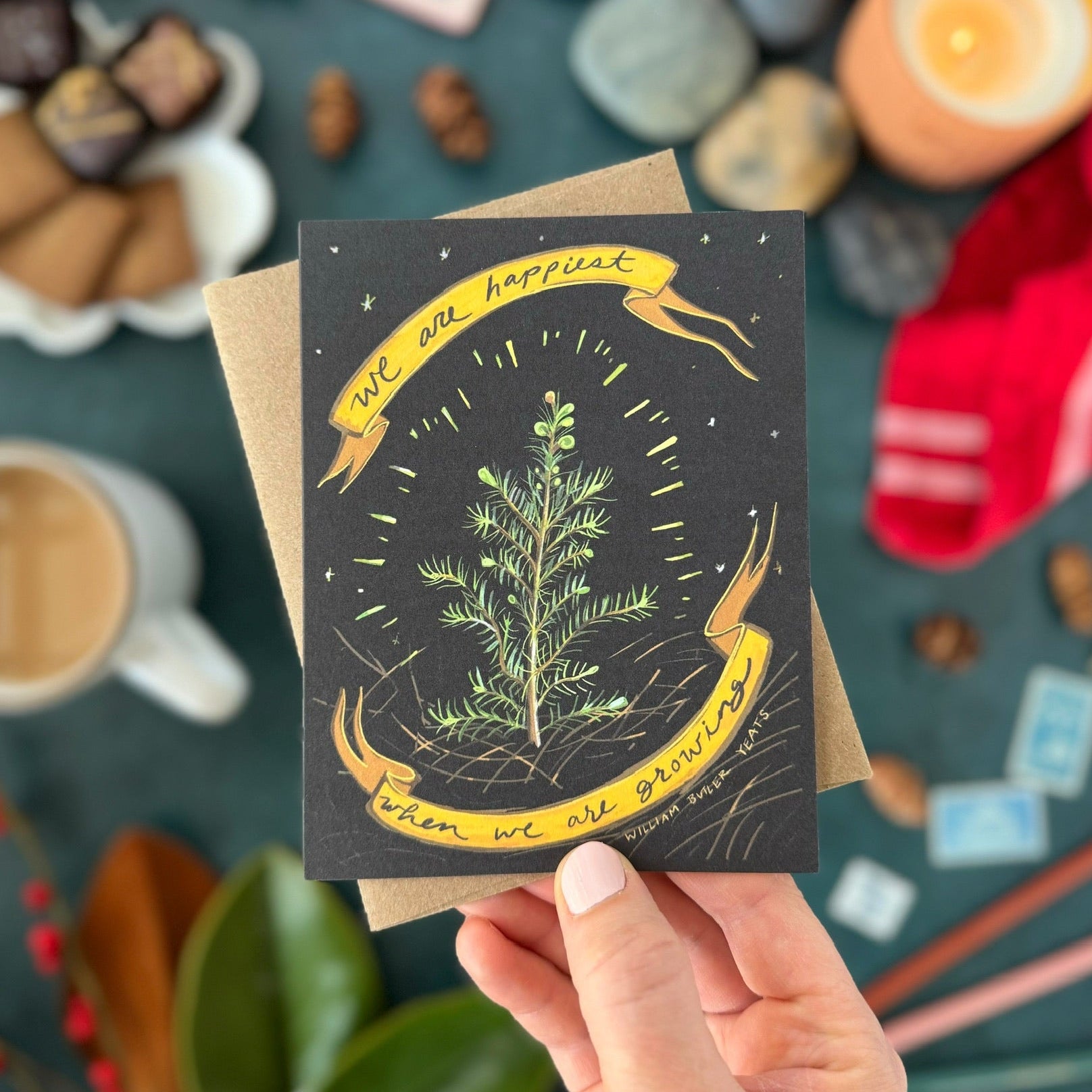 Hand holding a greeting card that says, "We are happiest when we are growing," with a plant illustration and text, surrounded by various items on a dark surface.