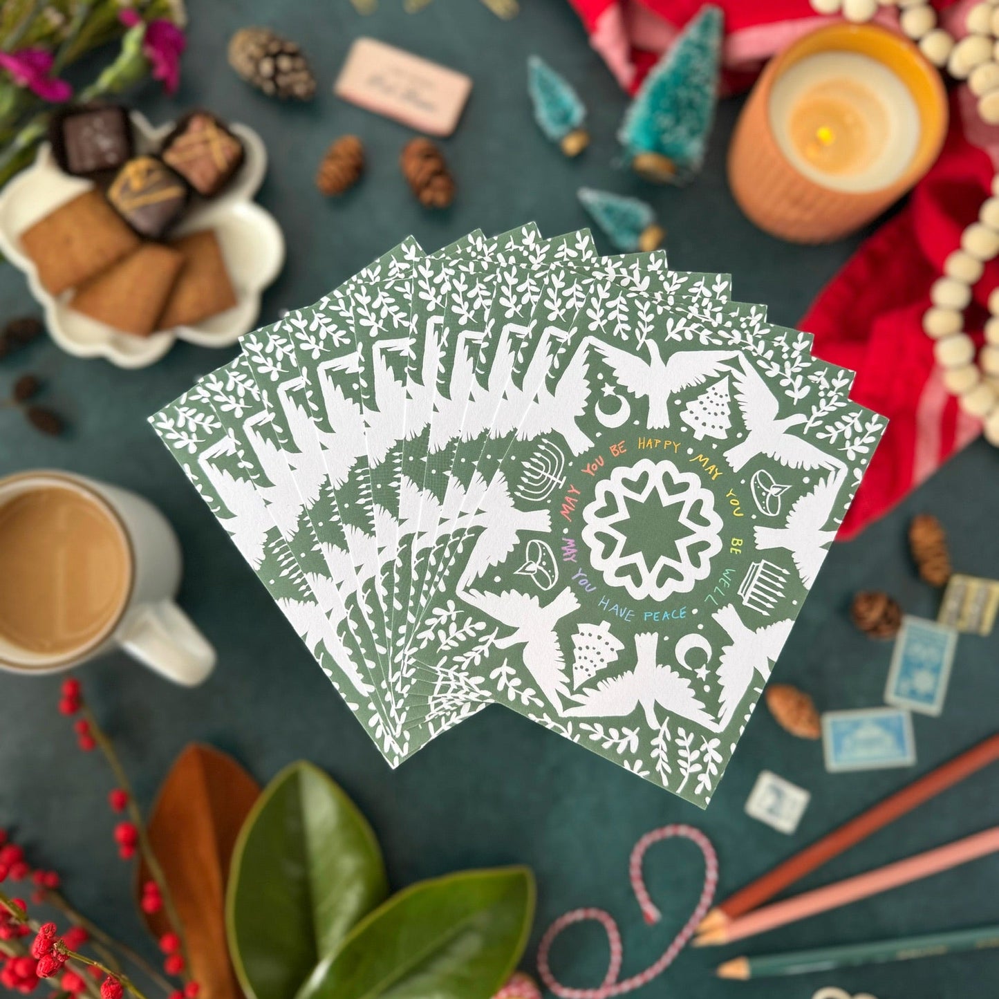 Green and white decorative postcards that say "May you be happy, may you be well, may you have peace," on a table with coffee, cookies, and candles.