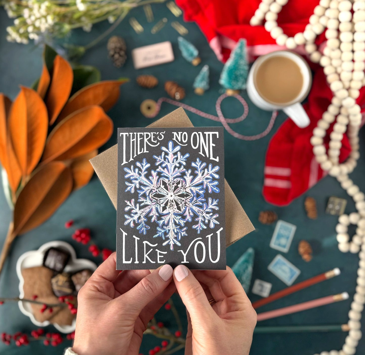 Hand holding a card with a snowflake design and text that says "there's no one like you," surrounded by festive items on a table.