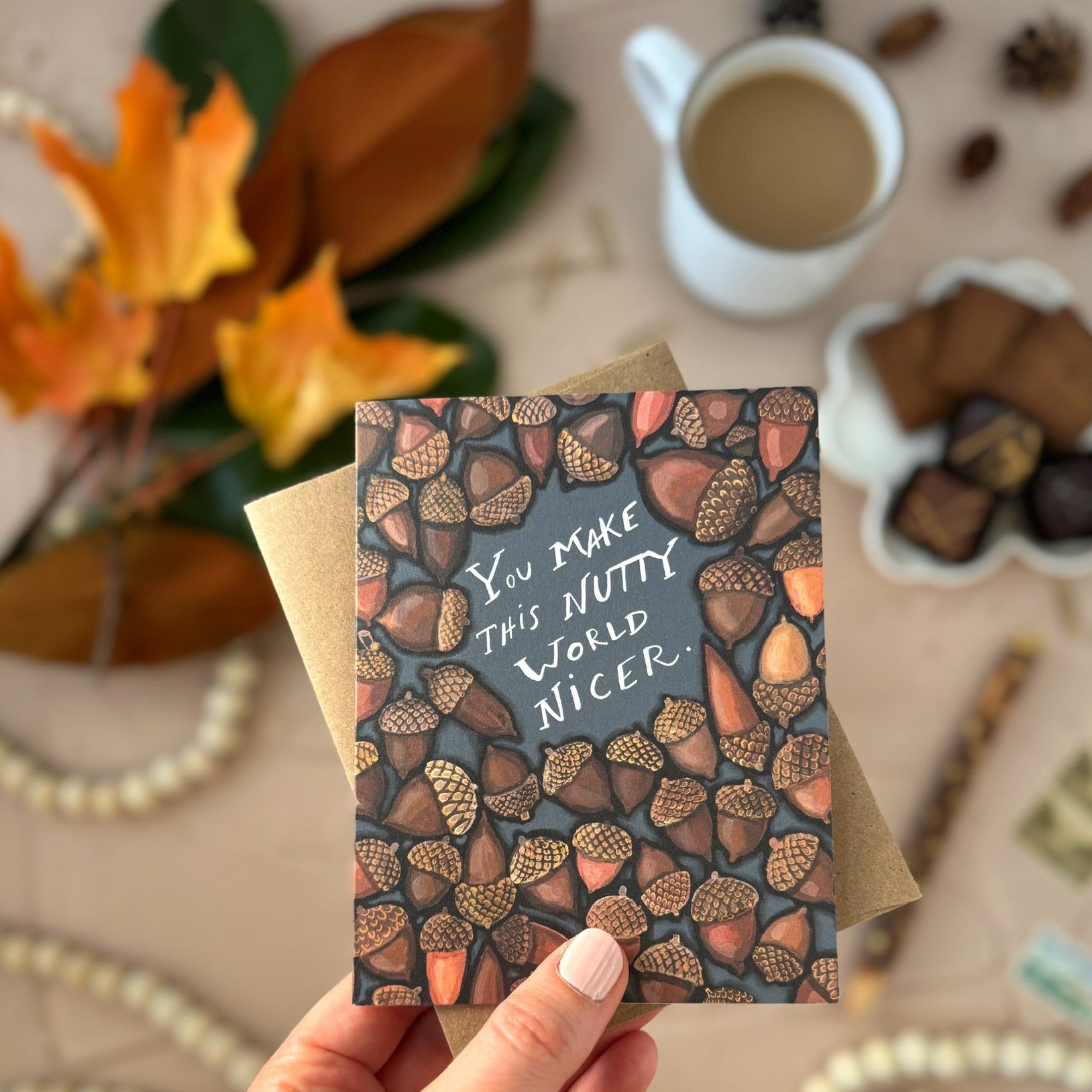 Hand holding a card that says, "You make this nutty world nicer," with acorn pattern and text, surrounded by autumn-themed decor.