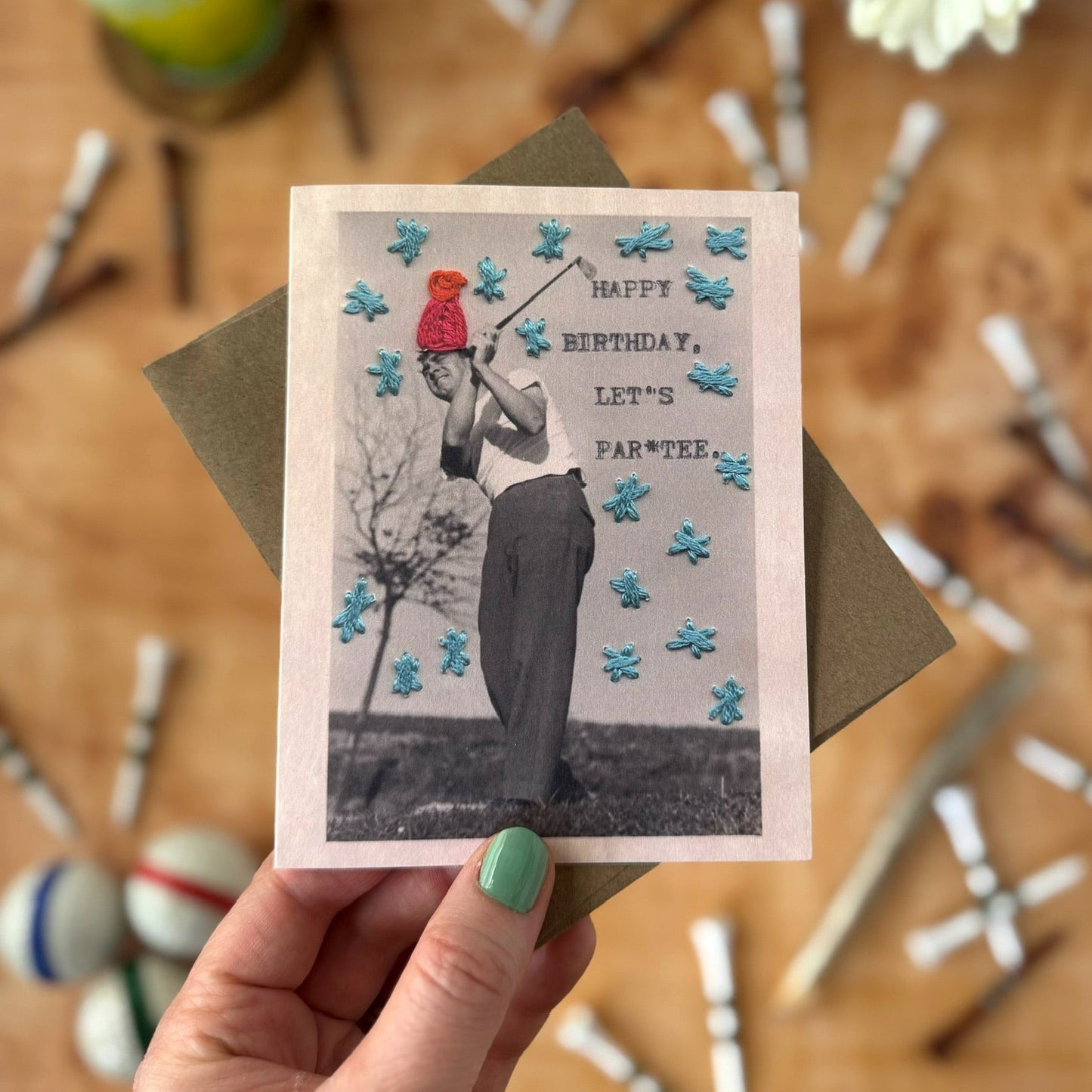 Hand holding a birthday card with a person in a red hat and white shirt golfing, surrounded by party supplies on a wooden surface.