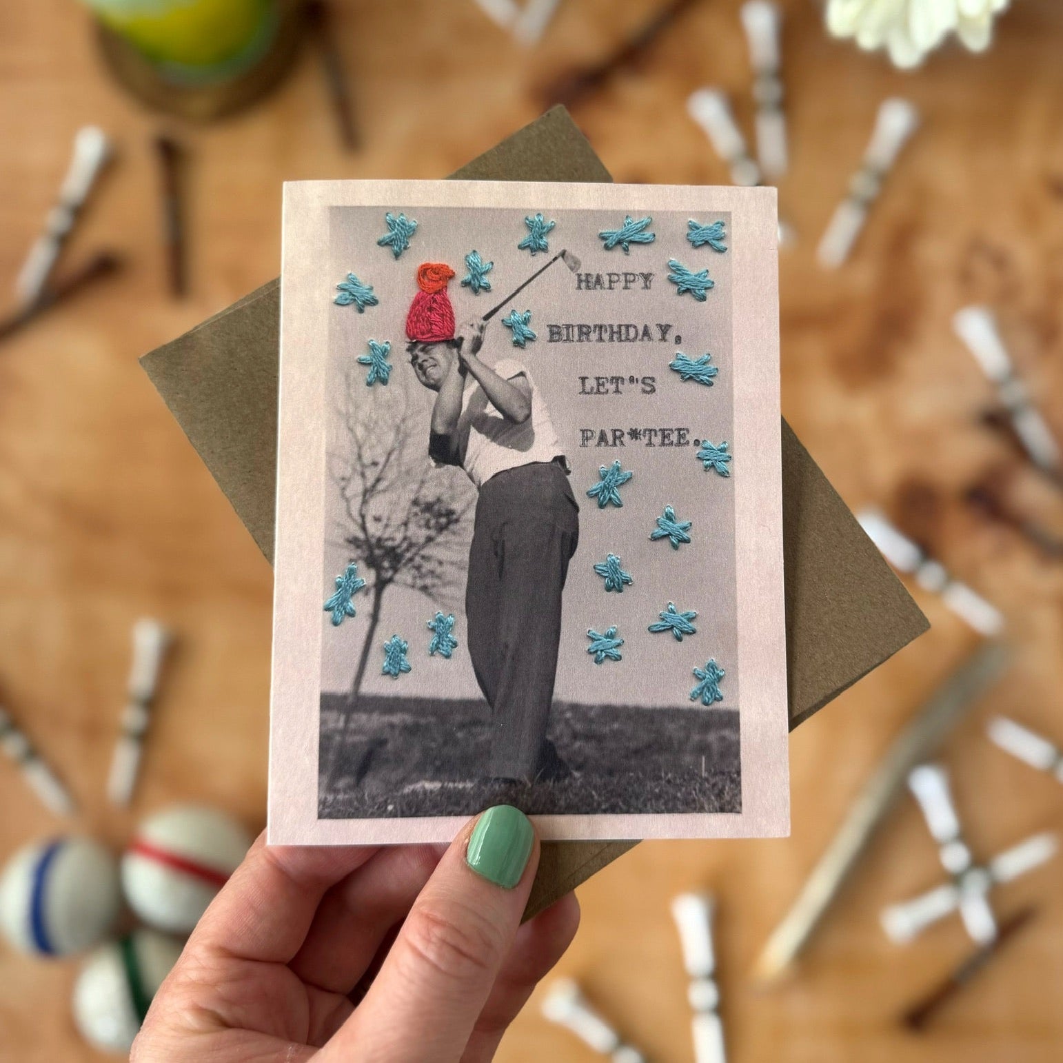 Hand holding a birthday card with a person in a red hat and white shirt golfing, surrounded by party supplies on a wooden surface.