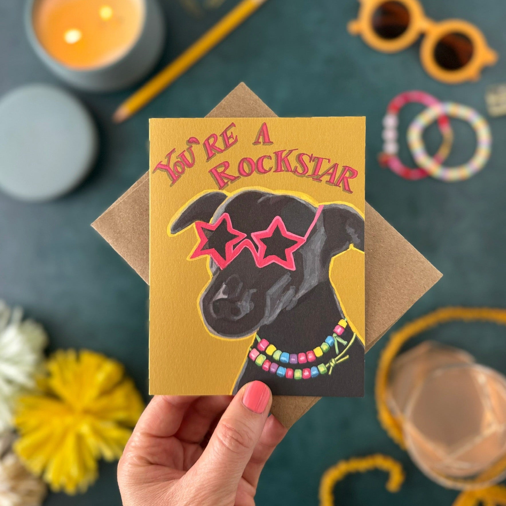 A hand holds a square greeting card featuring a black dog wearing pink star-shaped sunglasses and a colorful beaded necklace, with the text 'YOU'RE A ROCKSTAR' in pink. The card is shown with a brown envelope on a dark teal tabletop surrounded by styled props including a candle, yellow flowers, sunglasses, bracelets, and a pencil.