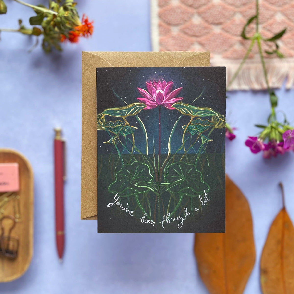 A greeting card with a kraft envelope, featuring an illustration of a pink lotus flower on dark green leaves against a black background, with the handwritten text 'You've been through a lot' at the bottom. The card is styled on a light blue surface with dried orange flowers, a red pen, a wooden tray with paperclips, and a pink patterned fabric swatch.