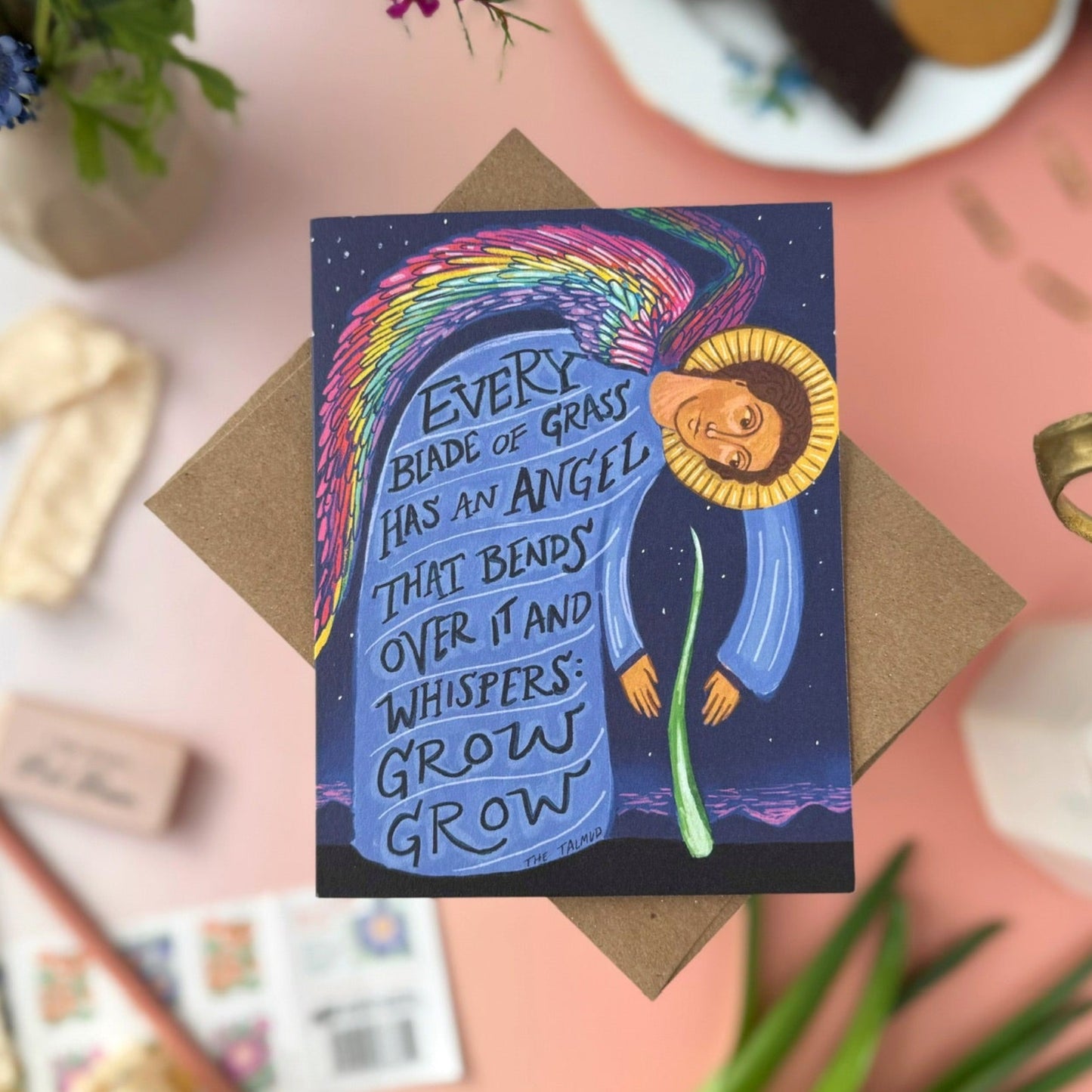 A card featuring a icon-like illustration of an angel with a blue body and yellow halo, surrounded by a rainbow wings and the text 'EVERY BLADE OF GRASS HAS AN ANGEL THAT BENDS OVER IT AND WHISPERS: GROW GROW'. The card is displayed on a kraft paper envelope on a pink background with decorative flat-lay props including flowers, a plate of cookies, and stamps.
