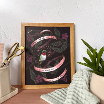 A framed art print featuring a hummingbird, pink banners with motivational text that says "May you faith be stronger than your fears", and green leaves on a dark background, styled on a wooden desk with a plant, scissors, and a textured towel.