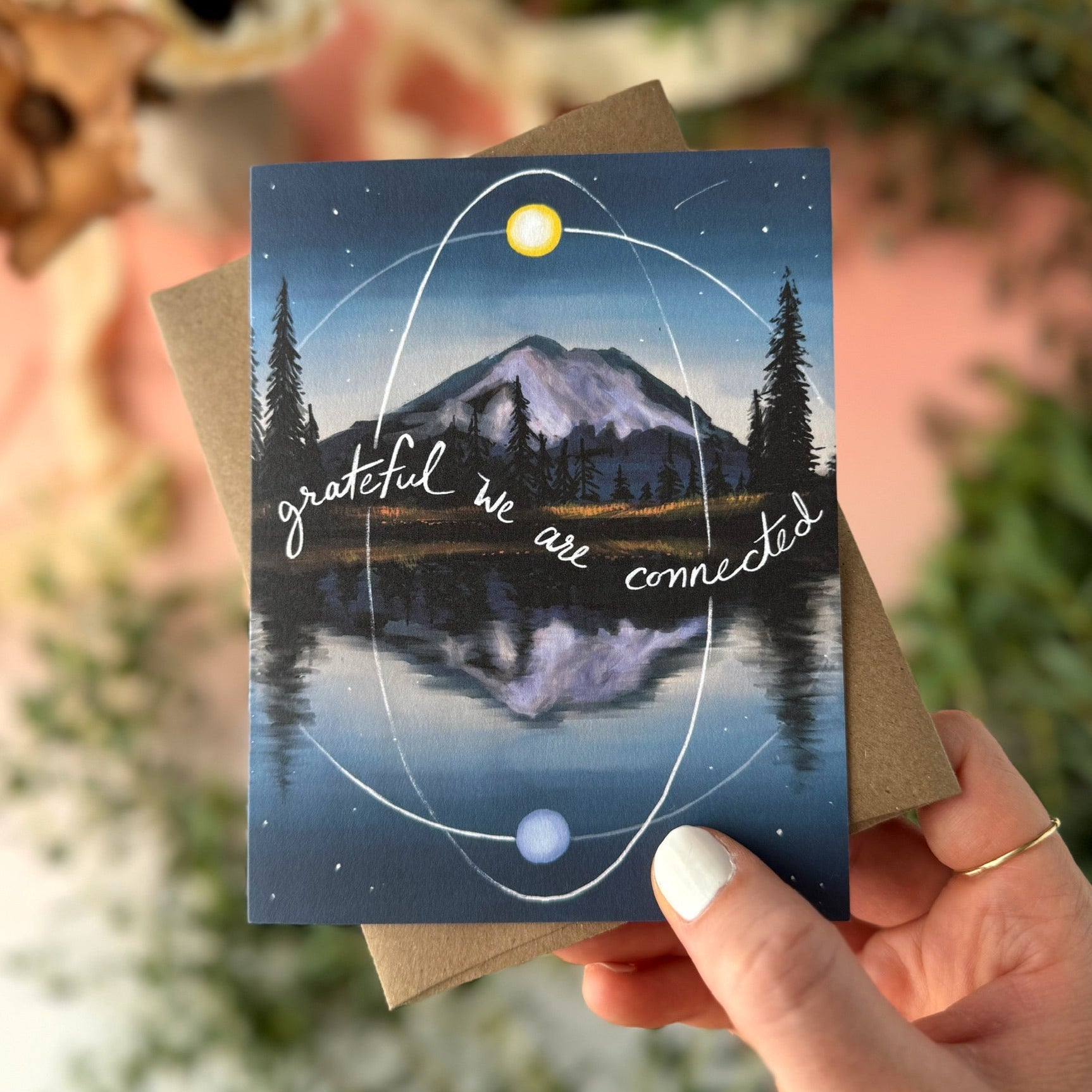 A hand holds a greeting card featuring a mountain reflected in a calm lake, with trees, stars, and white orbital lines and the phrase 'grateful we are connected.' The print is shown with a brown kraft envelope against a softly blurred peach and green background.