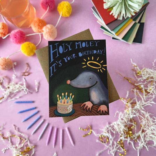 A flat-lay of a birthday card featuring a cartoon gray mole with a halo and a birthday cake, surrounded by party decorations like pom-poms, tinsel, and colored paper on a pink background. The card says "Holy Moley It's your birthday"