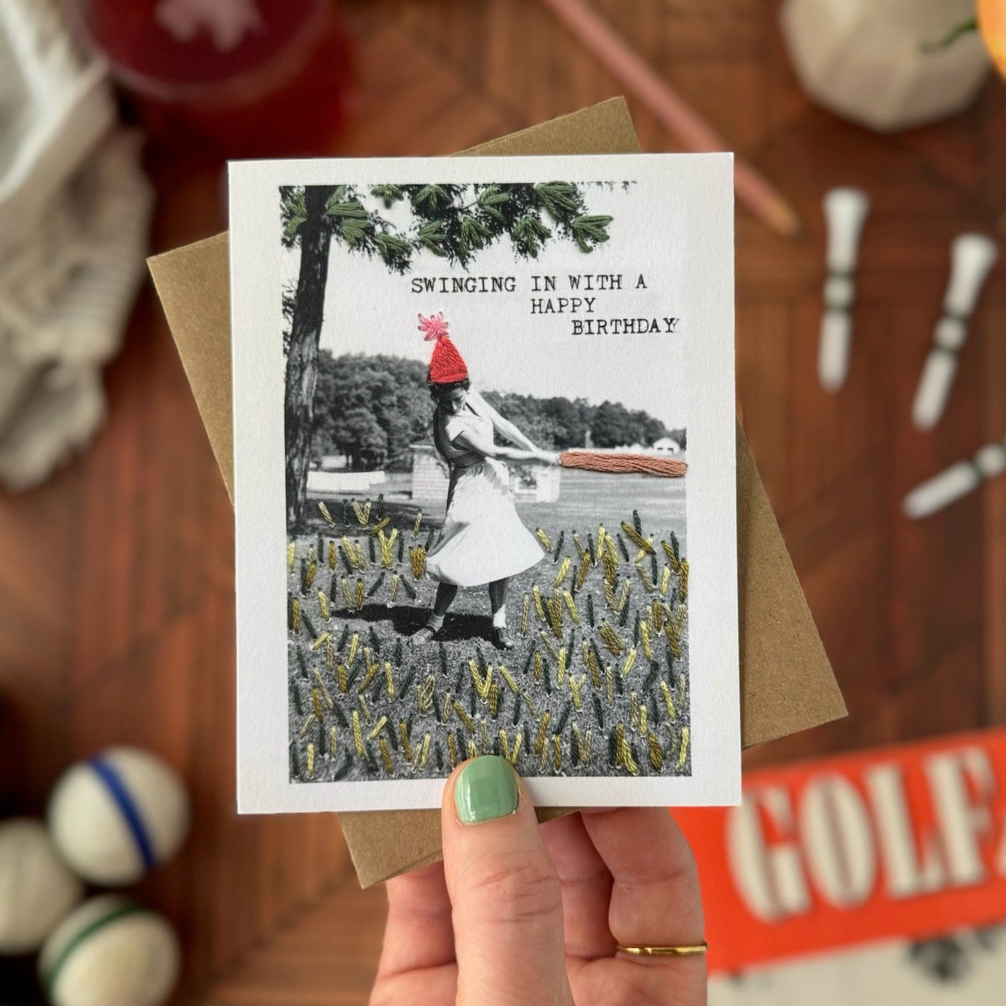 Hand holding a birthday card with a black and white photo of a lady  in a red hat and white dress golfing. The card is displayed against a blurred background with various objects.