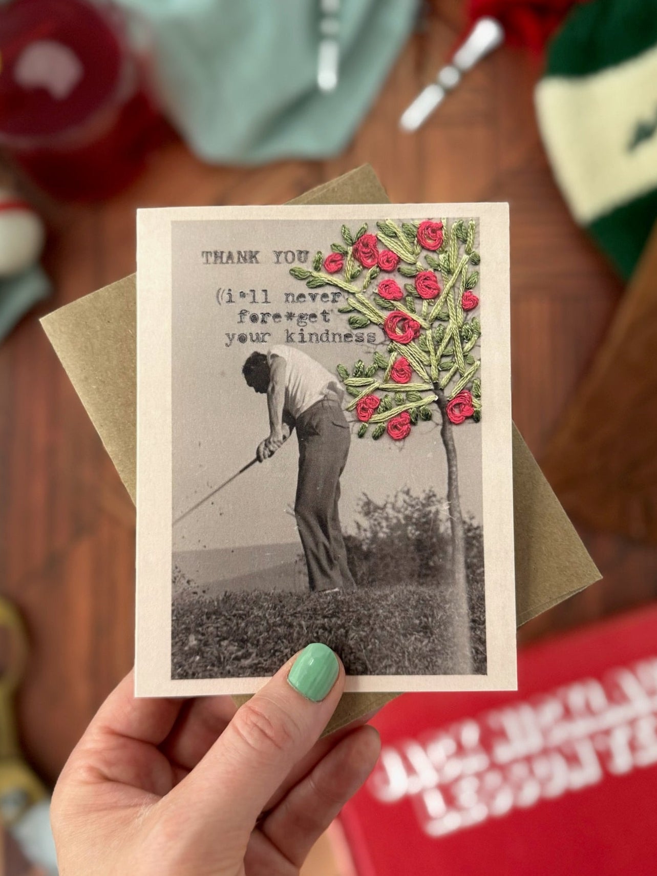 Hand holding a card with a vintage photo of a man golfing near a tree with flowers, surrounded by various objects on a wooden surface.