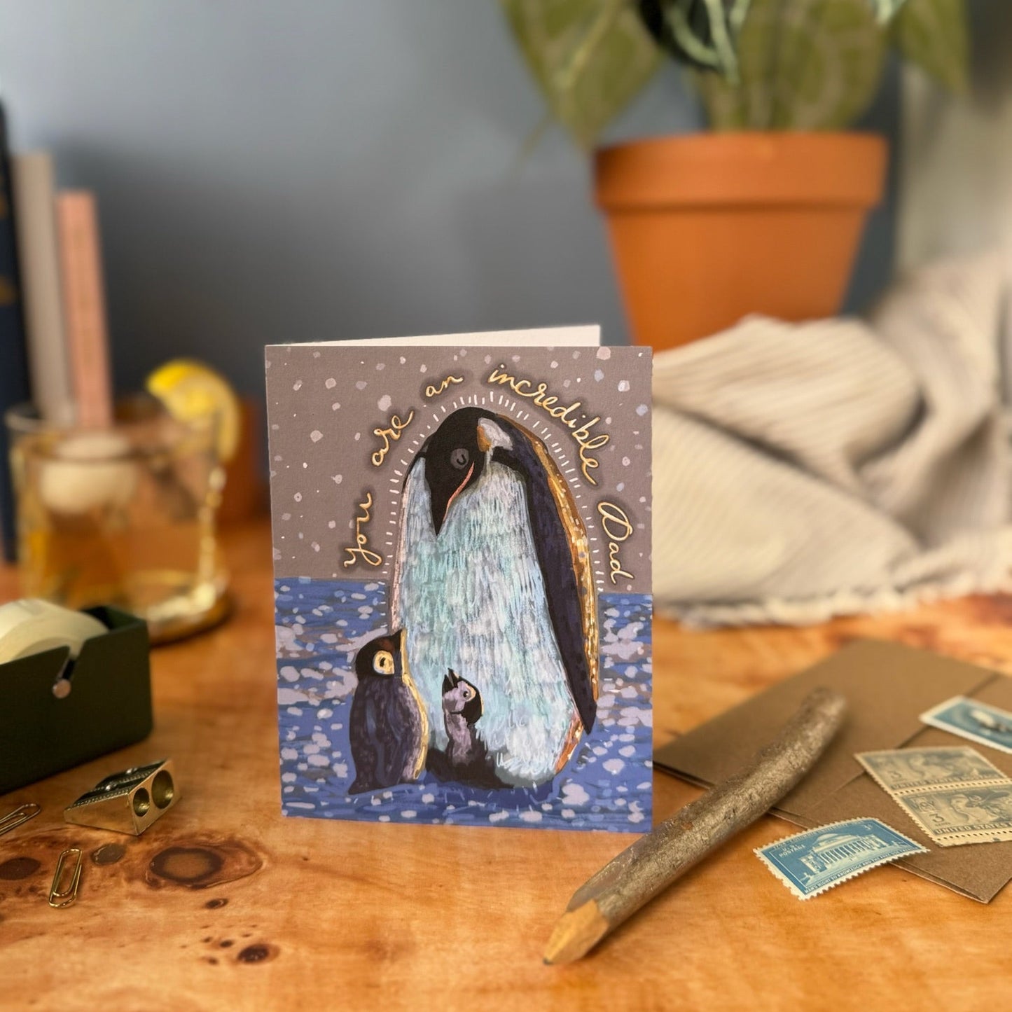Greeting card with penguin design on a wooden surface with plants in the background