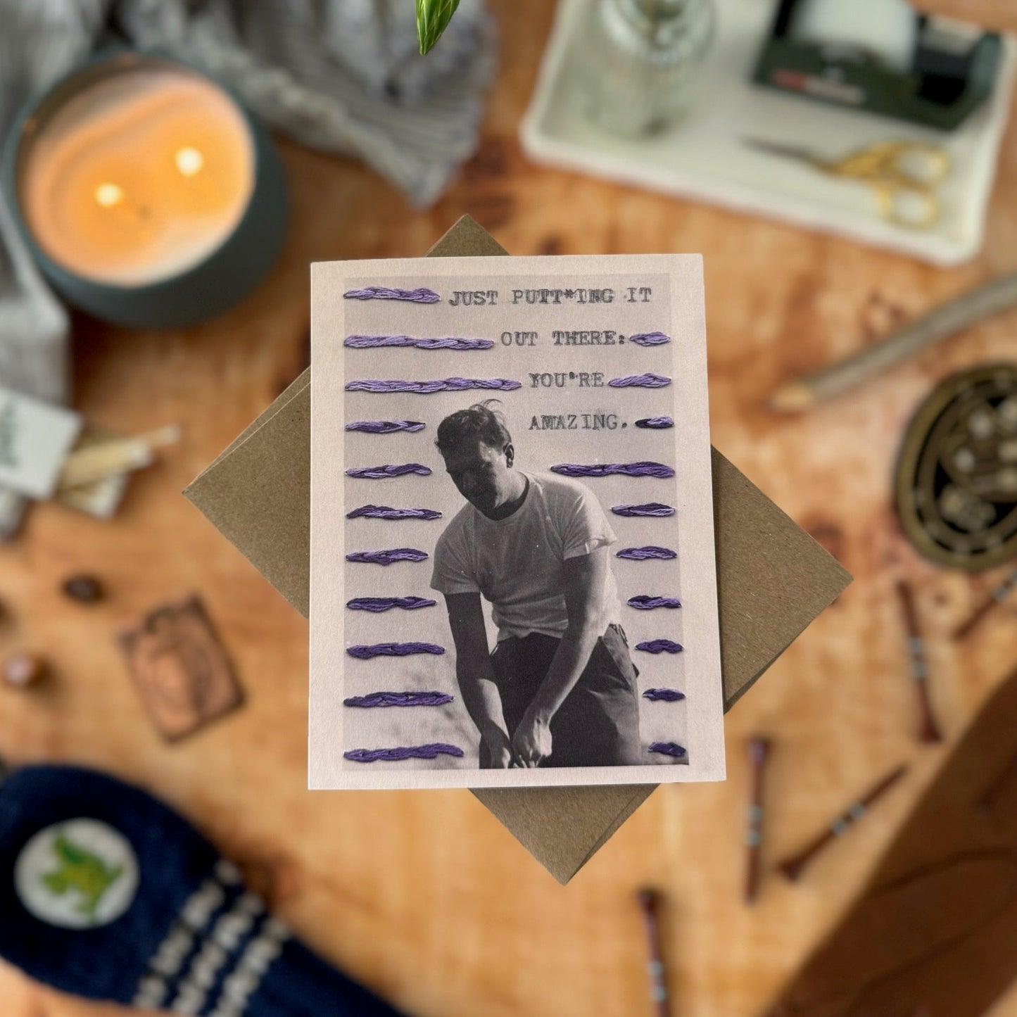 Greeting card with a printed image of a person golfing on a wooden surface with candles and stationery.