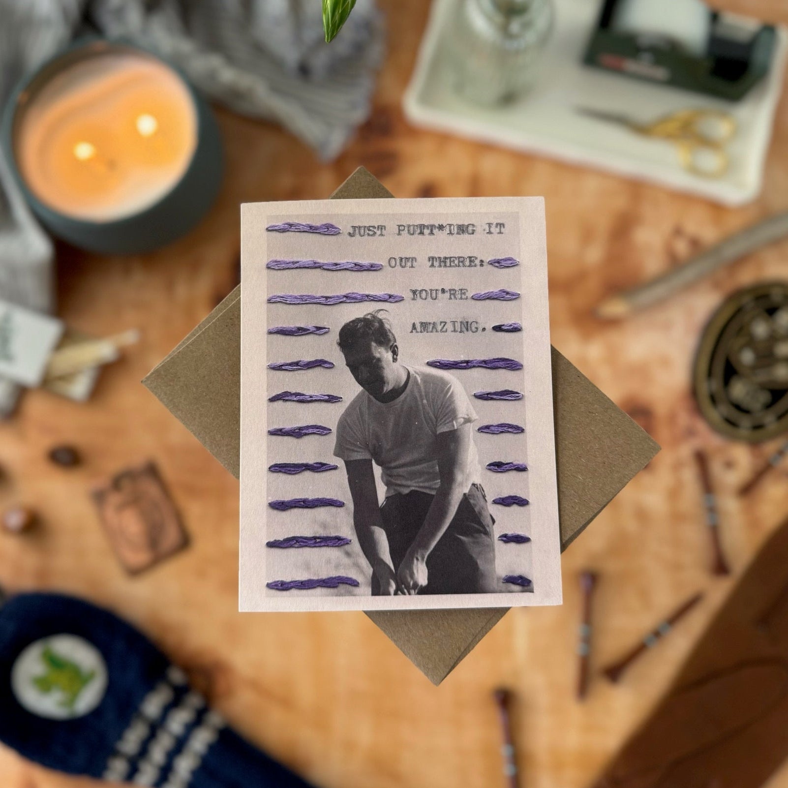 Greeting card with a printed image of a person golfing on a wooden surface with candles and stationery.