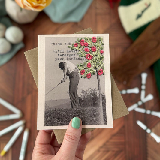 Hand holding a greeting card with a vintage-style illustration of a golfer and tree on a wooden surface.