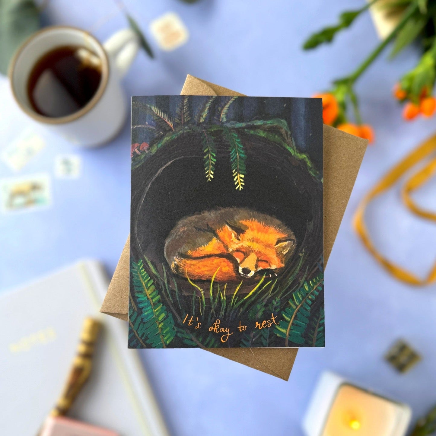 Greeting card with fox illustration on a blue surface with coffee and candles.