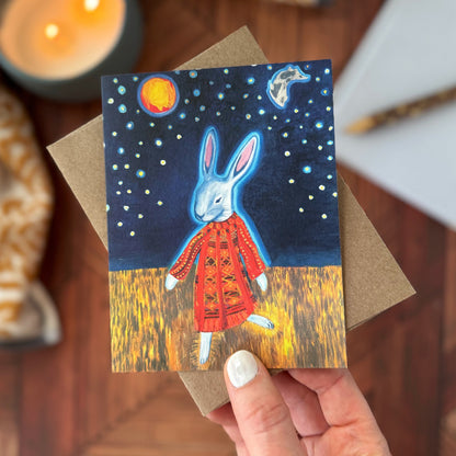 A hand holds a greeting card featuring an illustrated white rabbit in a quilted red coat under a starry night sky, with a brown envelope behind it and a warm wooden tabletop around it.