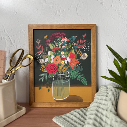 A framed floral art print featuring a colorful bouquet in a glass jar against a dark background, styled on a wooden desk with a plant, textured towel, and scissors in a cream holder against a textured white wall.