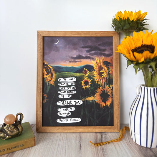 A framed art print featuring a sunset landscape with sunflowers and handwritten text, styled on a light wood surface next to a striped vase with sunflowers.
