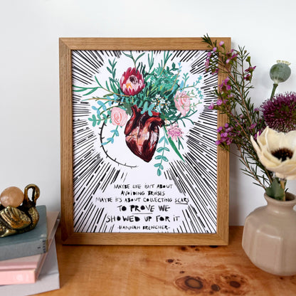 A framed art print featuring a watercolor anatomical heart surrounded by pink and red flowers and green leaves, with black hand-drawn rays and text, displayed on a wooden table with flowers and books.