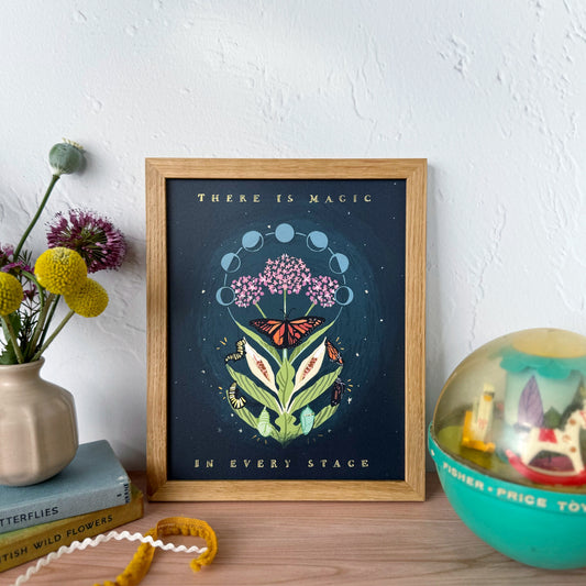A framed art print featuring a whimsical botanical illustration with butterflies and the text 'THERE IS MAGIC IN EVERY STAGE' displayed on a wooden tabletop next to a vase of flowers and a teal child's ball.