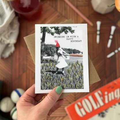 Hand holding a greeting card with a vintage-style photo of a woman golfing in a field, surrounded by a casual setting with a drink and flowers.