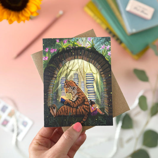 A hand holds an illustrated card featuring a tiger and girl reading among book stacks under a book arch, with a brown envelope behind it. The scene is styled on a pink surface with a sunflower, books, and greenery in the background.