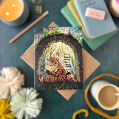 A greeting card featuring an illustrated tiger and girl reading among stacks of books under a book arch, displayed on a kraft envelope over a teal surface with books, a candle, flowers, and a cup around it.