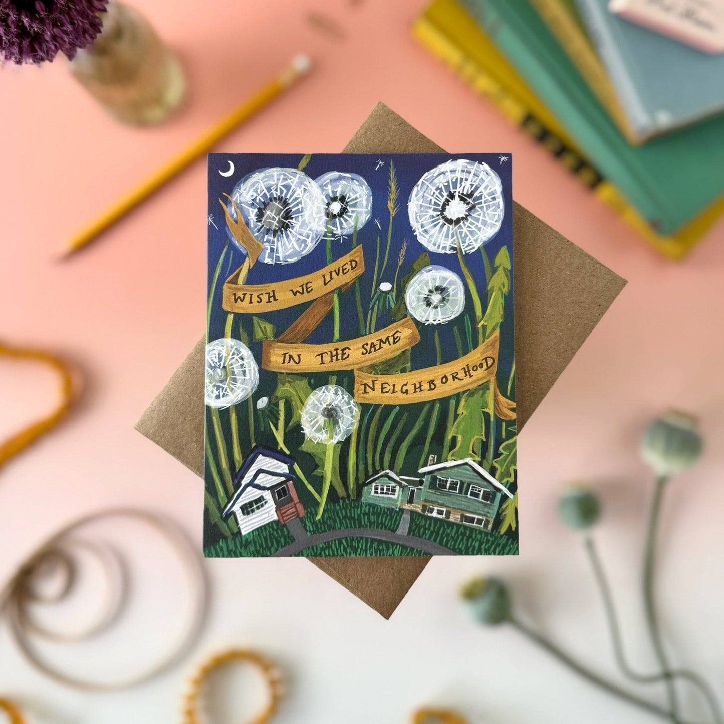 A square illustrated greeting card with a brown envelope, styled on a pink surface with books, a pencil, a purple flower, and decorative curls. The card artwork shows dandelions, houses, and a banner reading 'WISH WE LIVED IN THE SAME NEIGHBORHOOD'.