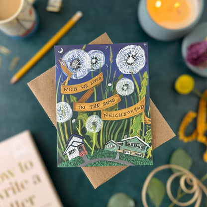 A square illustrated greeting card with a brown kraft envelope, photographed in a styled flat-lay scene. The card shows dandelions, small houses, and the text 'Wish we lived in the same neighborhood."