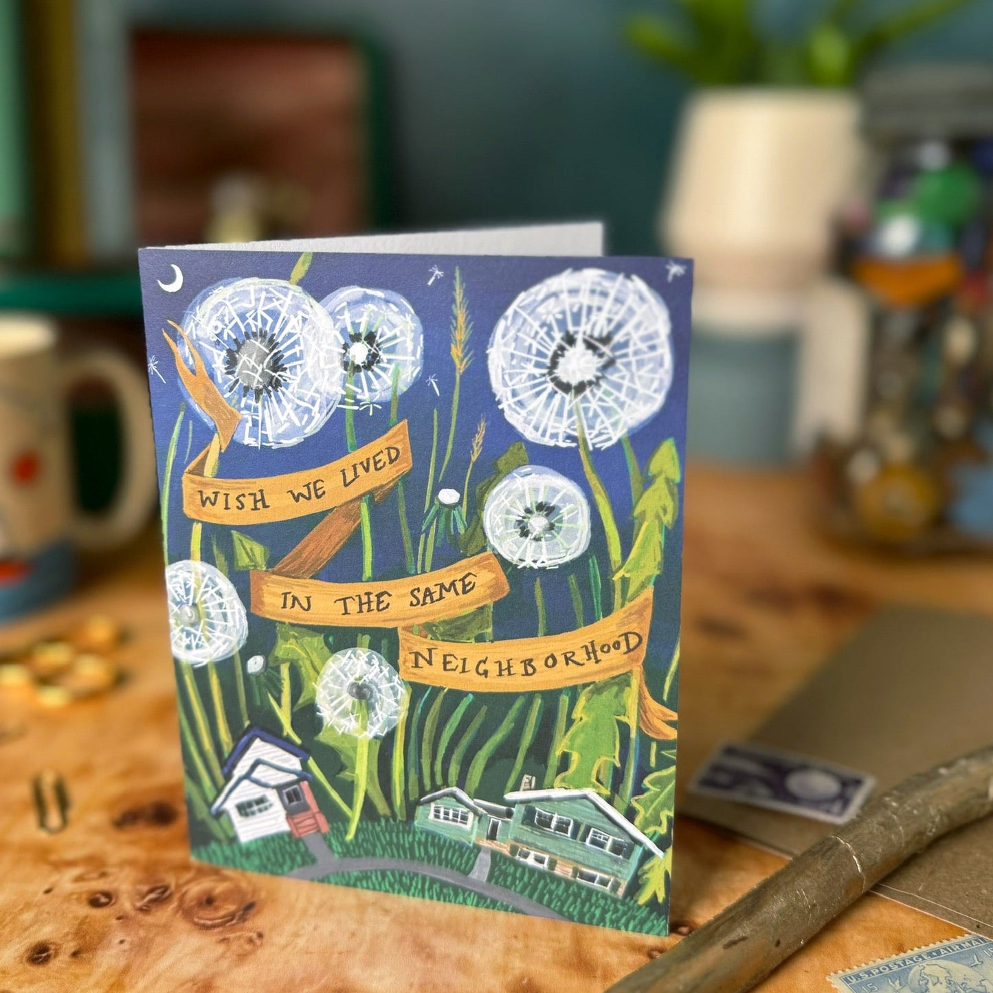 A square illustrated greeting card with a dandelion and house design, standing on a wooden tabletop in a softly blurred indoor setting.
The card says "Wish we lived in the same neighborhood."