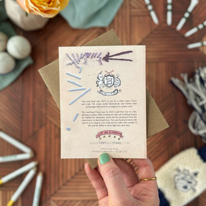 Hand holding a greeting card to show the embroidery detail on the back 
with branding on a wooden surface.