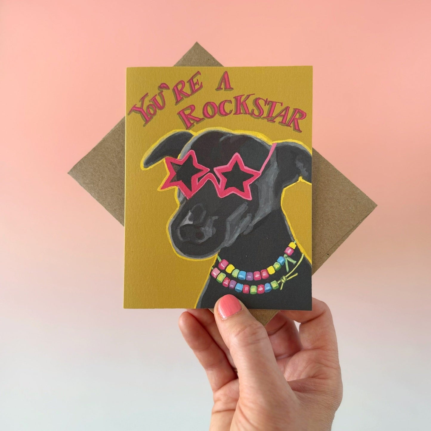 A hand holds a greeting card featuring a black dog wearing pink star-shaped sunglasses and a colorful beaded necklace on a mustard yellow background, with the text 'You're a Rockstar.' A brown envelope is partially visible behind the card against a soft pink-to-blue gradient background.