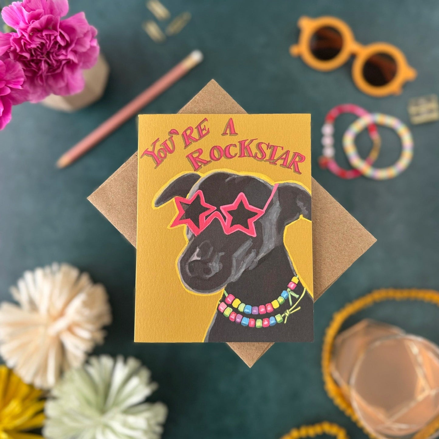 A  greeting card with a kraft envelope, styled in a flat lay on a dark teal background. The card features a black dog wearing pink star-shaped sunglasses and a colorful beaded necklace, with the text 'YOU'RE A ROCKSTAR' in pink.