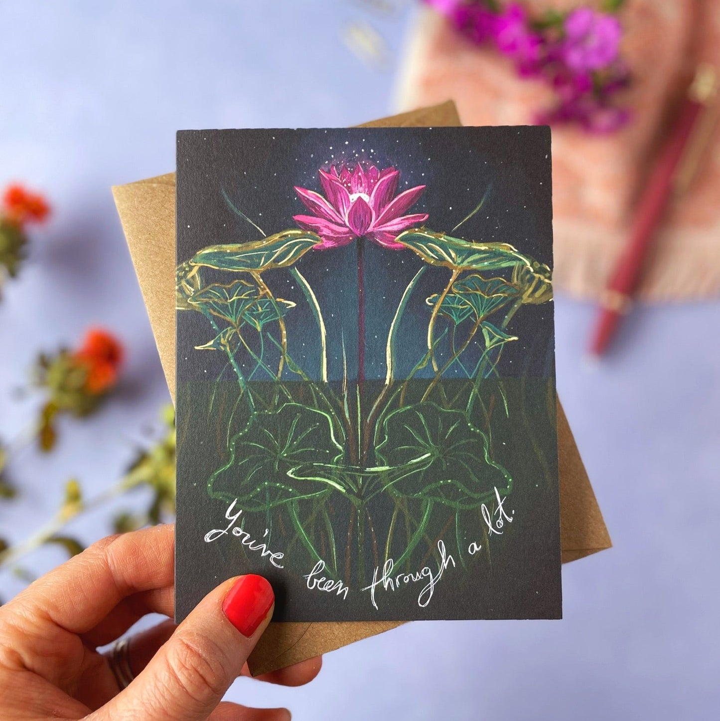 Hand holding a card with a pink lotus flower design and text against a blurred floral background