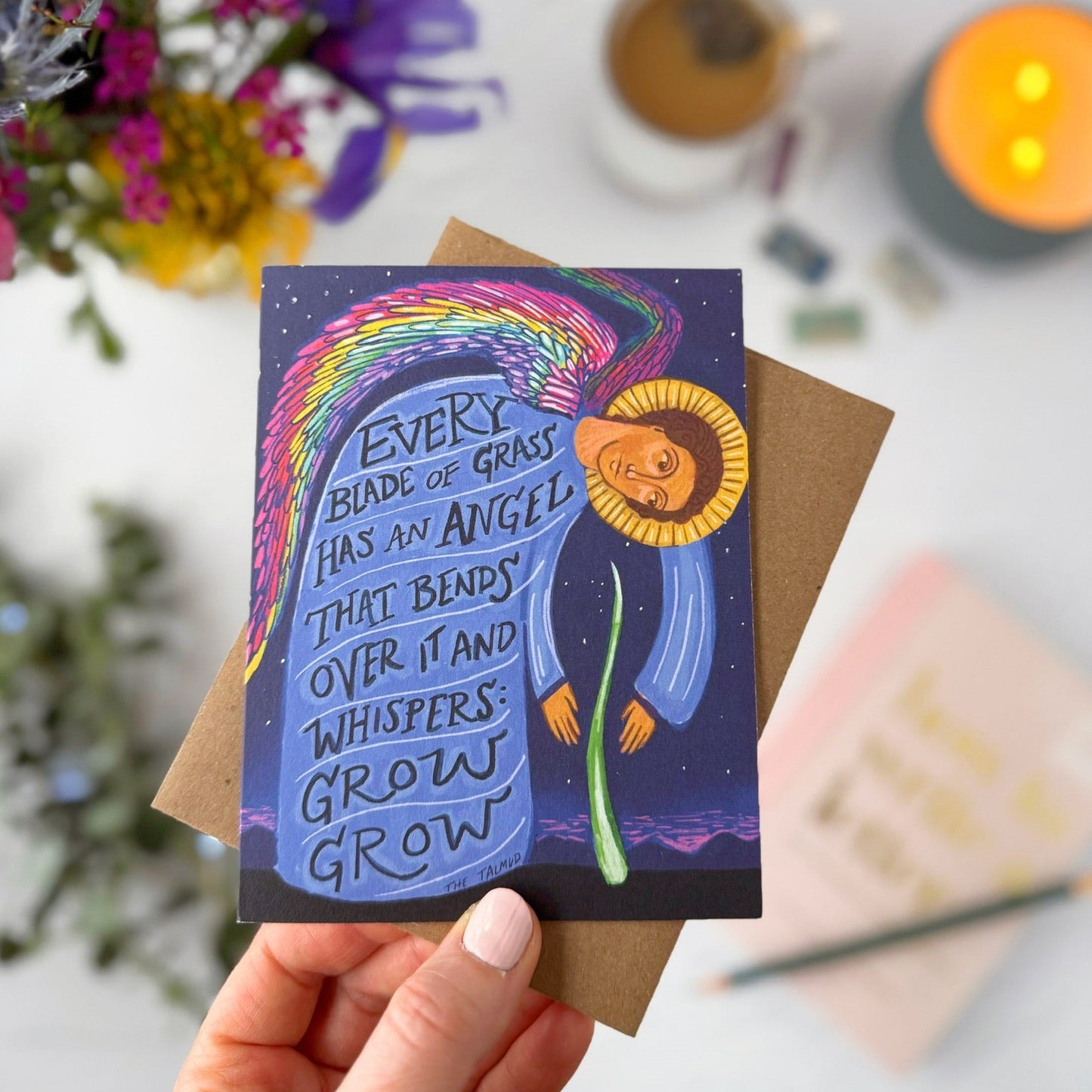 Hand holding a card with a colorful illustration and text, surrounded by flowers and candles.