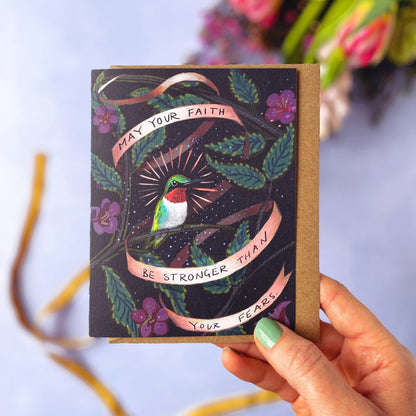 Hand holding a card with a colorful design of a hummingbird and text that says "May your faith be stronger than your fears", with flowers in the background.