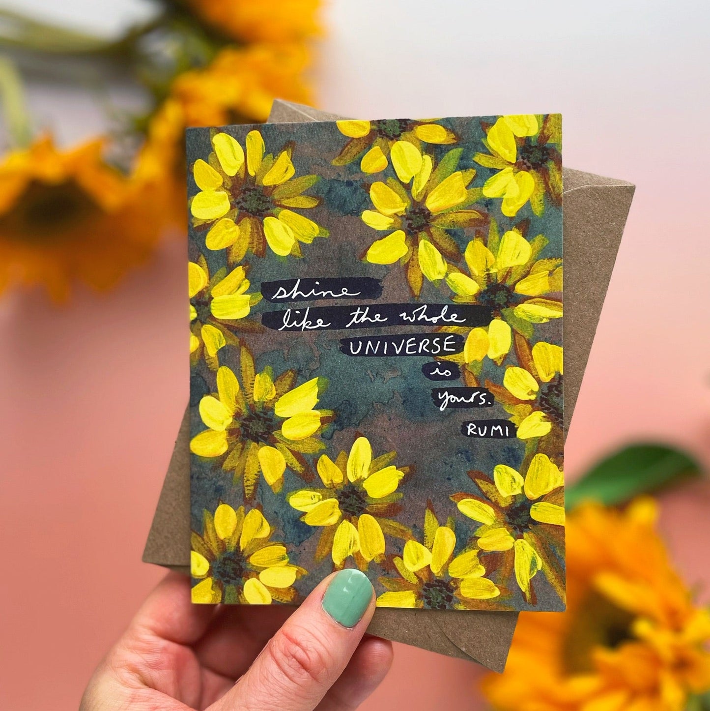 Card with yellow flowers and text held by a hand, surrounded by sunflower-like flowers.