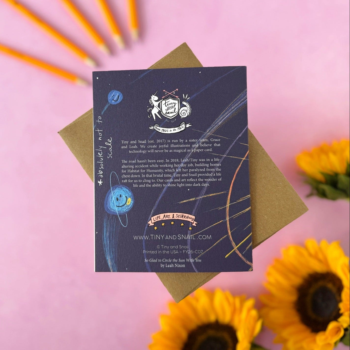 A flat-lay photograph of the back of the "so glad to circle the sun with you" card from 'Tiny and Snail' on a kraft envelope, set against a pink background with decorative yellow sunflowers and pencils.